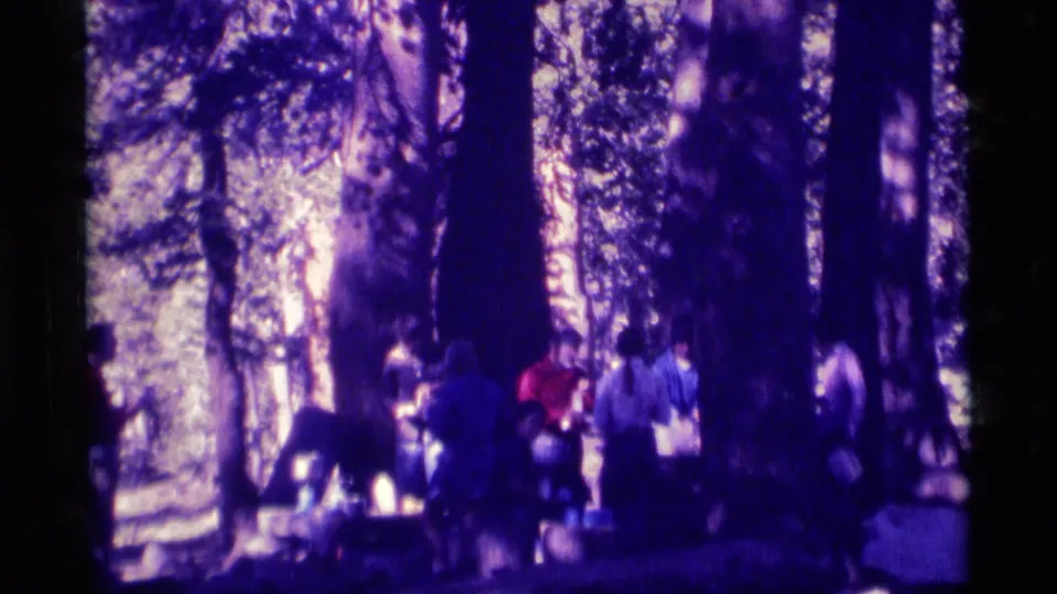 1975 8mm frame shows adults in a forested Minaret Wilderness campsite area, some near gear or a possible communal table.