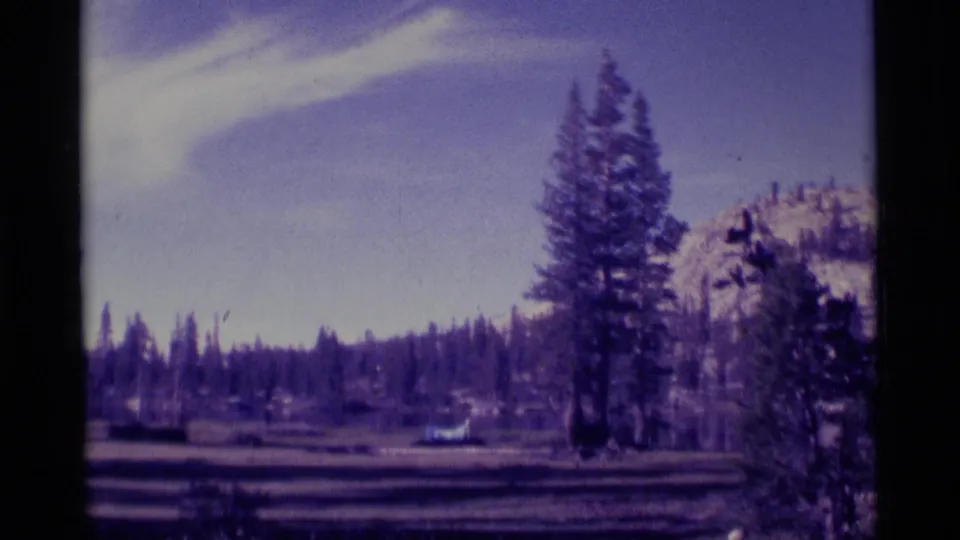 1975 8mm Minaret Wilderness frame showing an open clearing, tall pines, distant forest, and snow-dusted mountains.