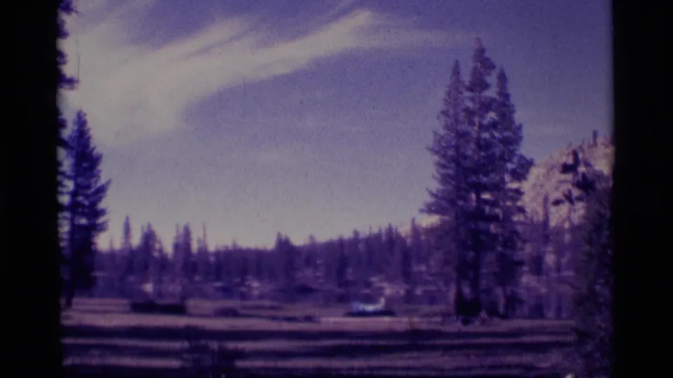 1975 Minaret Wilderness home-movie frame with a wide forest clearing, conifers, distant snow-dusted mountains, and dark 8mm frame edges.