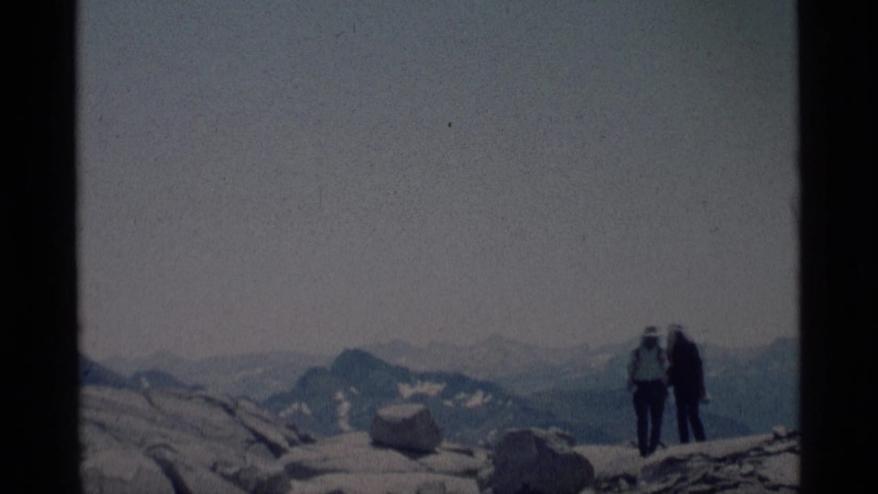 Two hikers with backpacks stand on rocky terrain with distant mountain peaks behind them.
