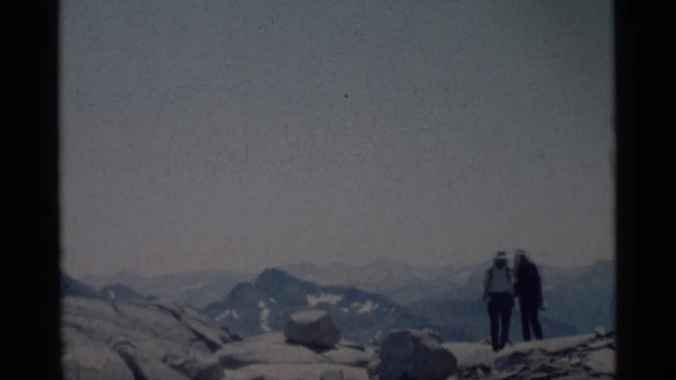 1975 8mm Minaret Wilderness footage showing two backpacked hikers on a rocky summit with hazy, snow-patched mountains in the distance.