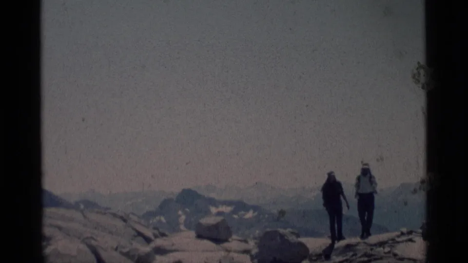1975 home-movie frame of two backpacked hikers crossing open rock in California mountain wilderness with distant ridges behind them.