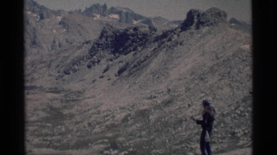 1975 8mm frame from Minaret Wilderness showing a lone hiker paused on a rocky alpine slope below jagged mountains.