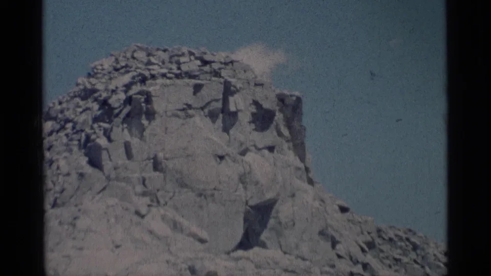1975 8mm frame from Minaret Wilderness showing a rugged, fractured mountain cliff rising against a pale blue sky.