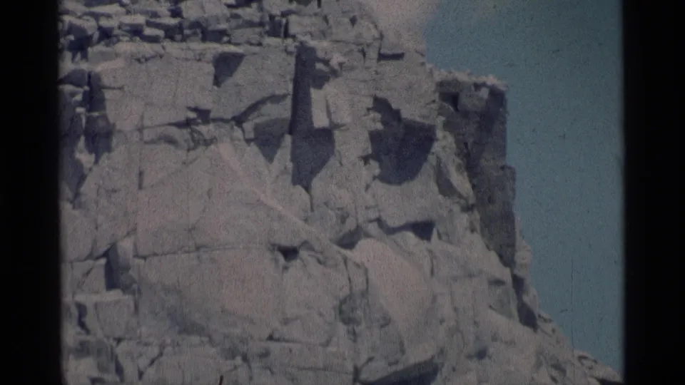 1975 8mm frame from Minaret Wilderness showing a massive angular cliff section with layered fractures and a narrow band of blue sky.