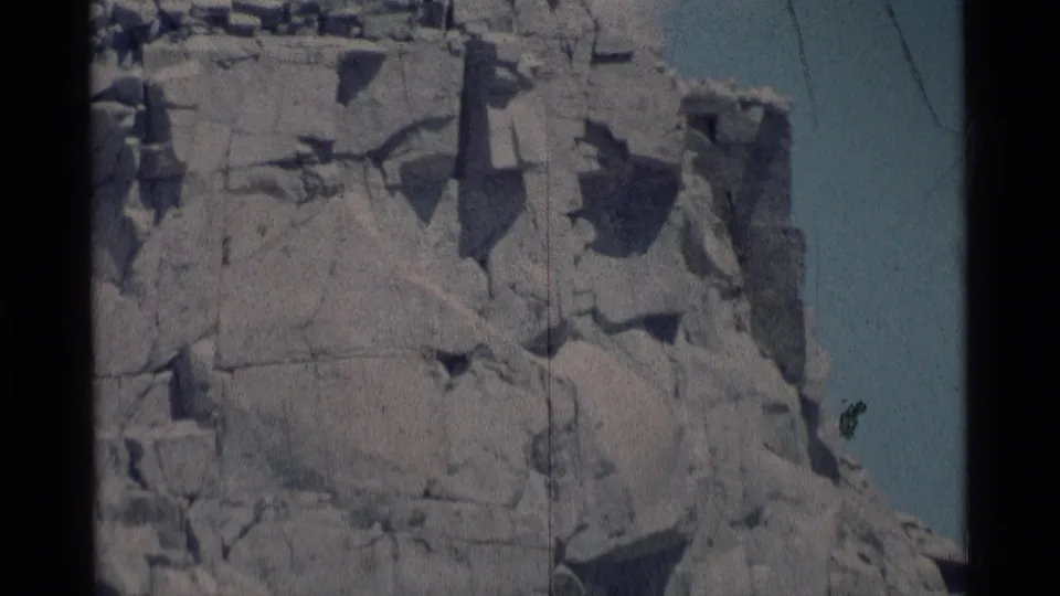 1975 8mm frame from Minaret Wilderness showing the jagged cliff face, pale sky, and a visible vertical film scratch near the right side.