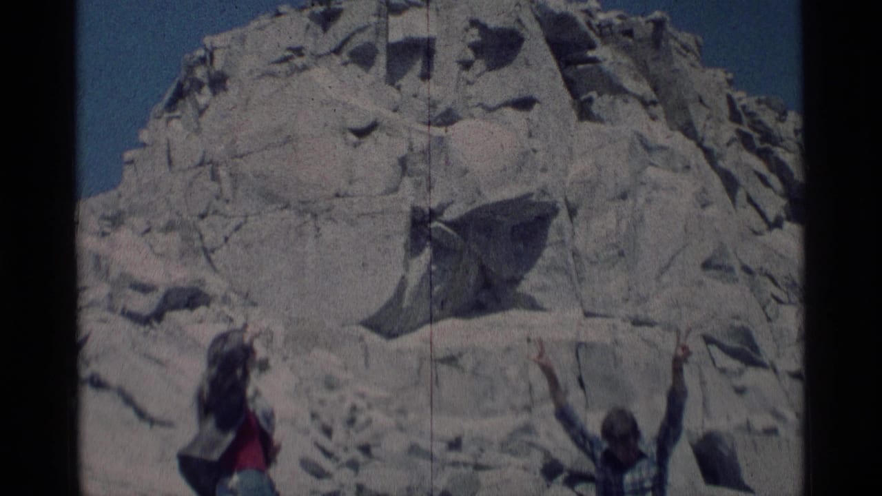 Two people stand below a pale granite rock formation under blue sky.