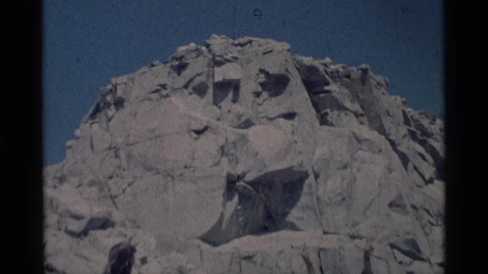 Minaret Wilderness home-movie frame with a sculpted granite face dominating the view and two small figures cropped near the lower edge.