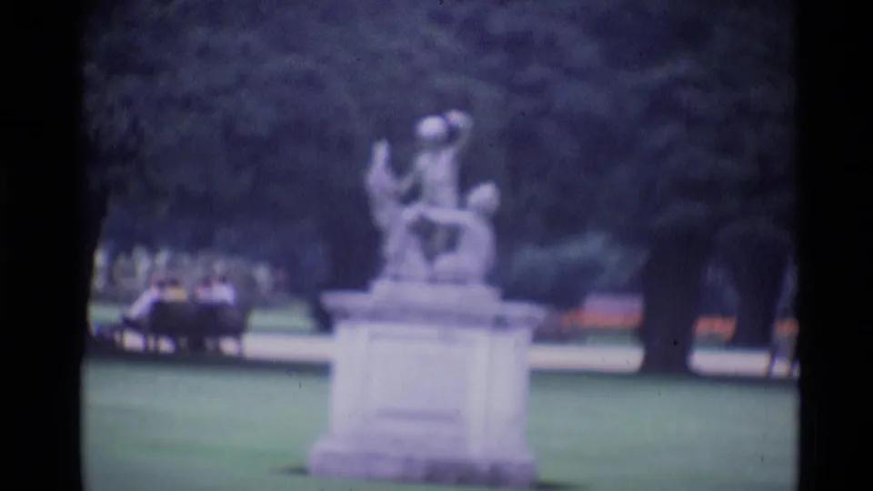 1969 London park frame with a pale classical statue on a pedestal, lawn, trees and a small vehicle in the background.