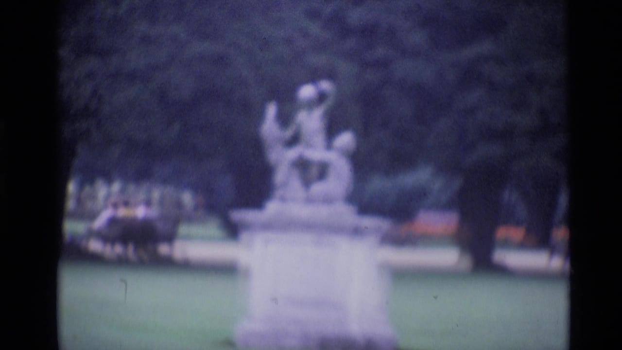 1969 London 8mm frame showing a blurred stone garden statue, surrounding lawn, trees, and dark film edges.