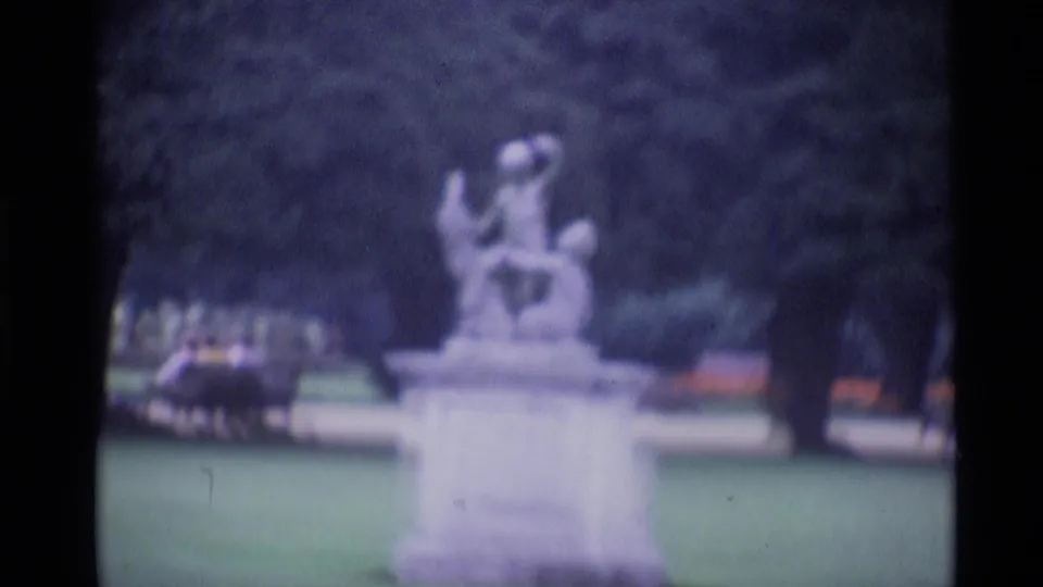 1969 London 8mm frame showing a blurred stone garden statue, surrounding lawn, trees, and dark film edges.