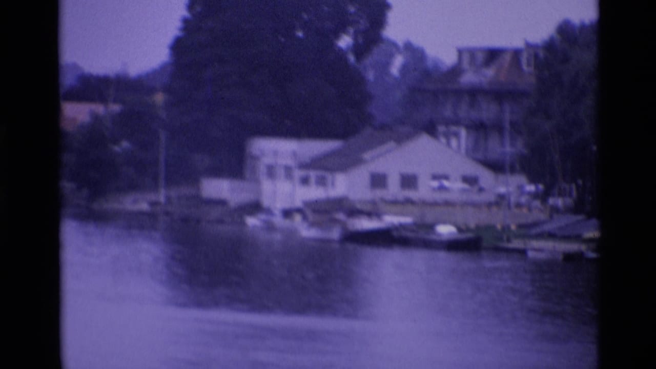 1969 London riverside view with a white waterside building, small boats, trees and calm water in a purple-tinted 8mm frame.