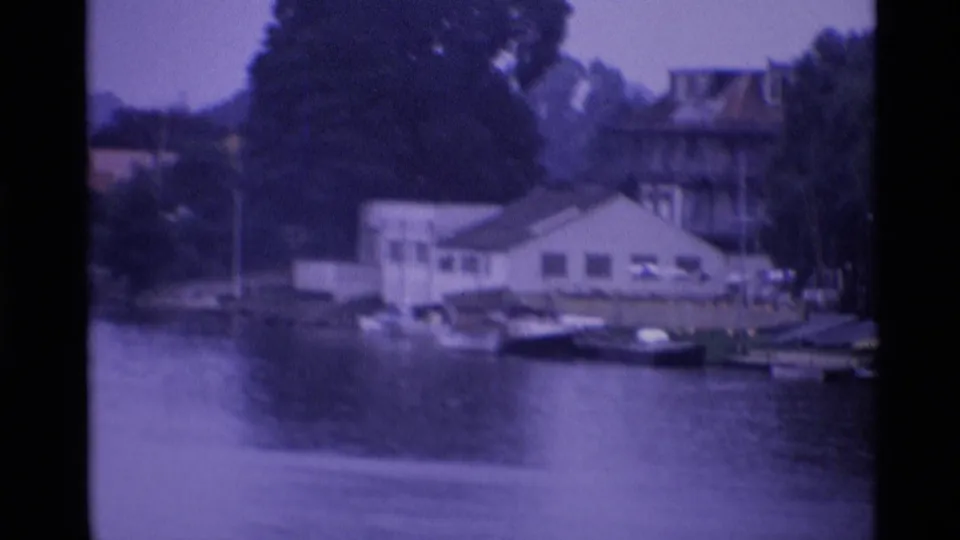 1969 London riverside view with a white waterside building, small boats, trees and calm water in a purple-tinted 8mm frame.