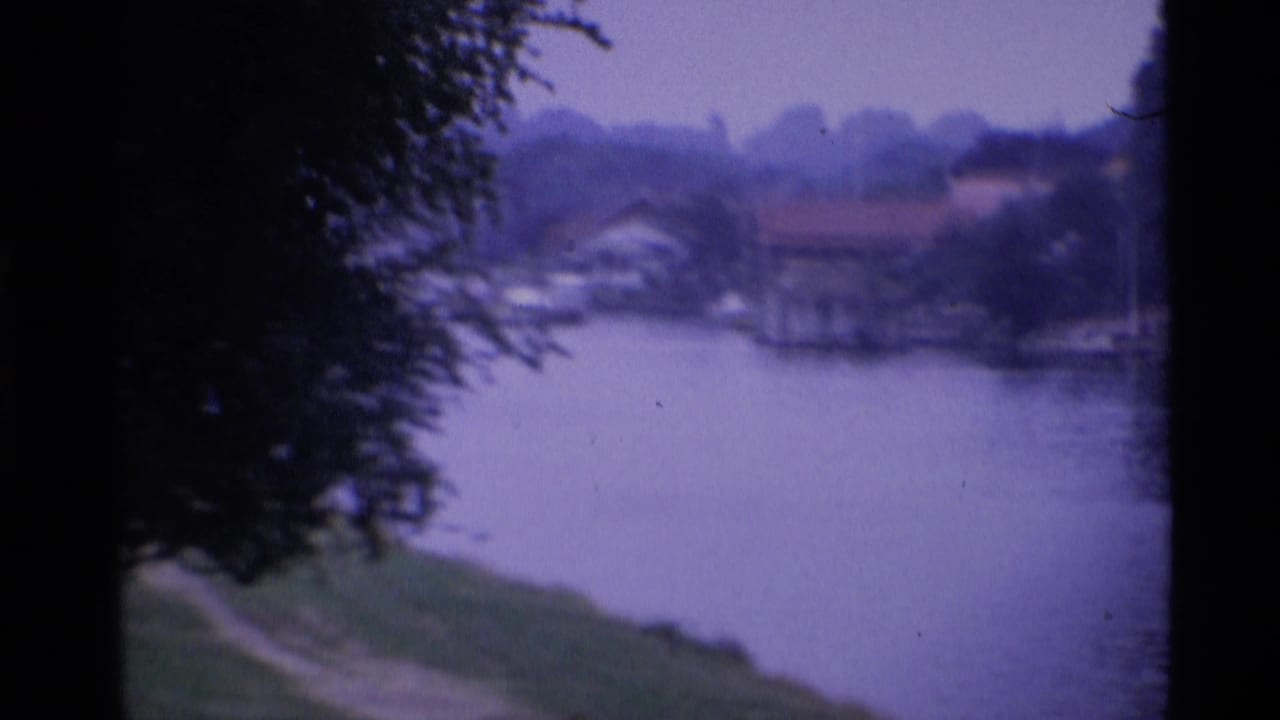 1969 London 8mm frame showing a calm river, grassy near bank, trees, and distant waterside homes under a purple cast.