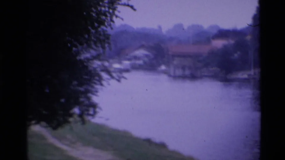 1969 London 8mm frame showing a calm river, grassy near bank, trees, and distant waterside homes under a purple cast.