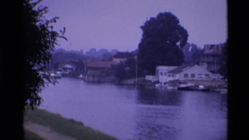 1969 London riverside frame with houses and moored boats across the river, framed by dark trees and film borders.