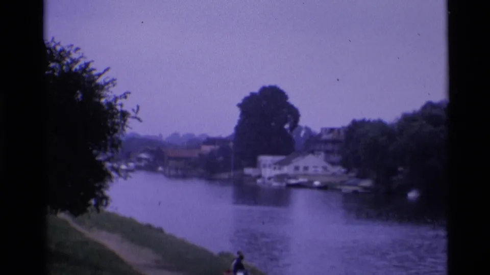 1969 London 8mm frame showing riverside houses, moored boats, trees, and a distant person on the near bank.