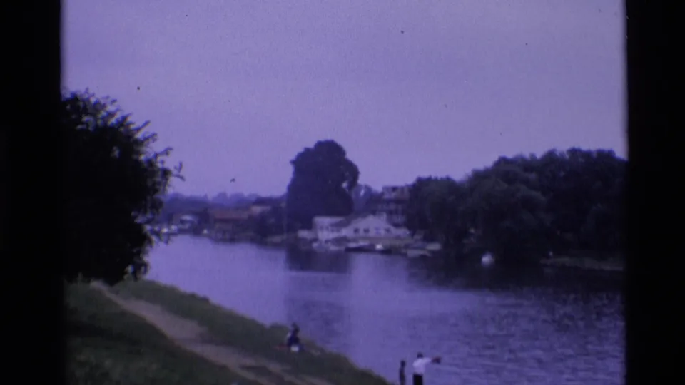 1969 London home-movie frame with three distant people by the riverbank, waterside houses, boats, and heavy tree cover.