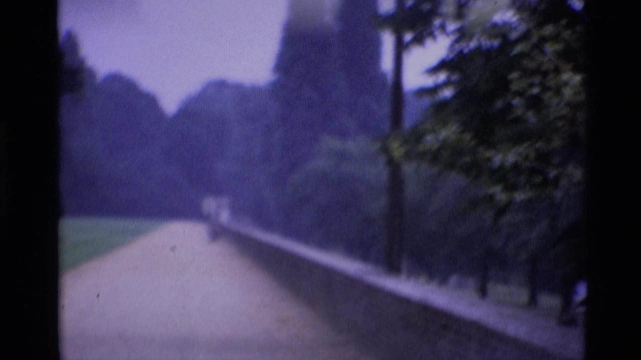 A London park path runs beside a low brick wall and trees, with a small distant visitor visible under a pale cloudy sky.