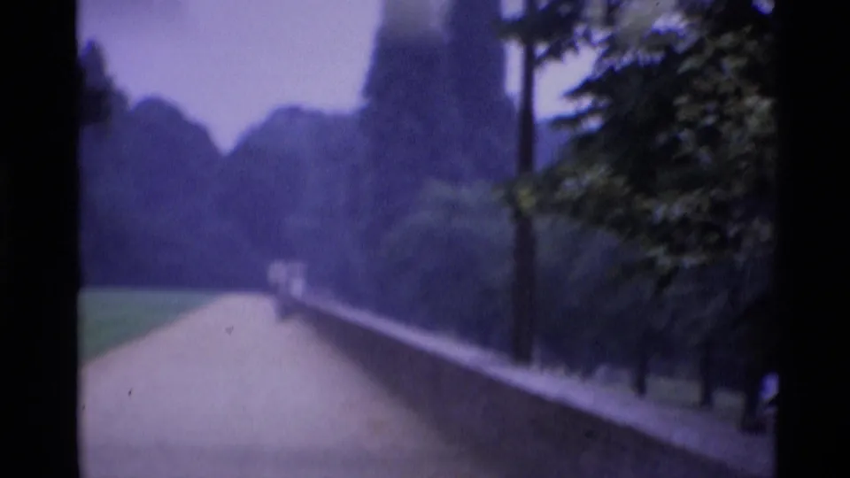 A London park path runs beside a low brick wall and trees, with a small distant visitor visible under a pale cloudy sky.