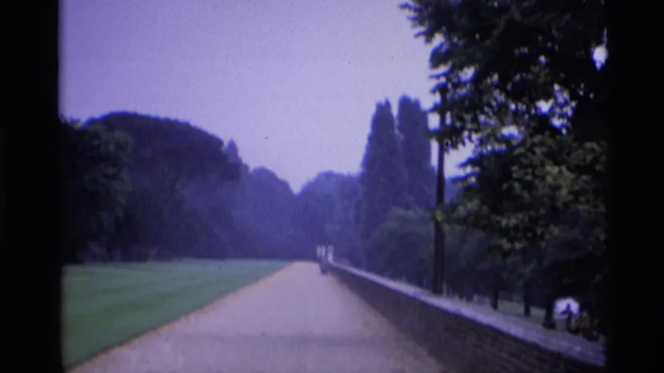 The 8mm frame shows a broad London park walkway, green lawn, mature trees, a low brick wall, and distant walkers near the path.