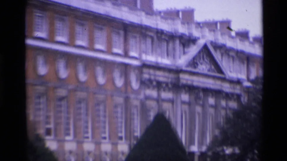 Closer 1969 London home-movie frame showing the red brick facade, classical trim, and a clipped hedge in the garden foreground.