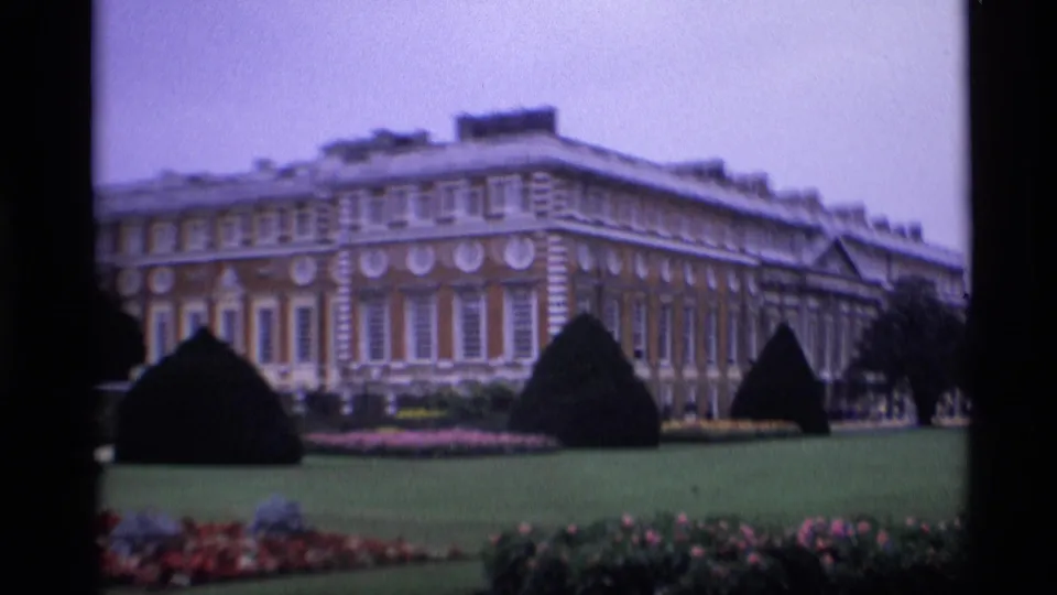 Wider 1969 London 8mm frame of a grand red brick building viewed across a formal lawn, hedges, and flowerbeds.