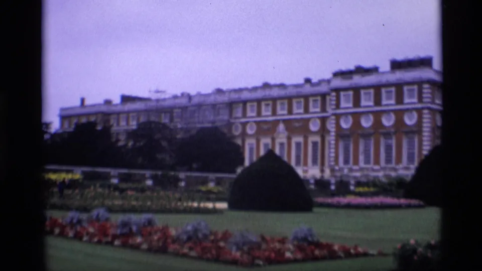 Wide 1969 London home-movie view of a formal garden foreground and grand red brick building facade beyond it.
