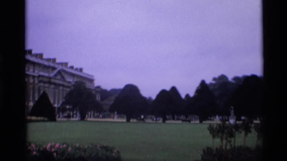 1969 London home-movie frame showing a formal green lawn, clipped trees, flowerbeds, and a partial view of a grand classical building.