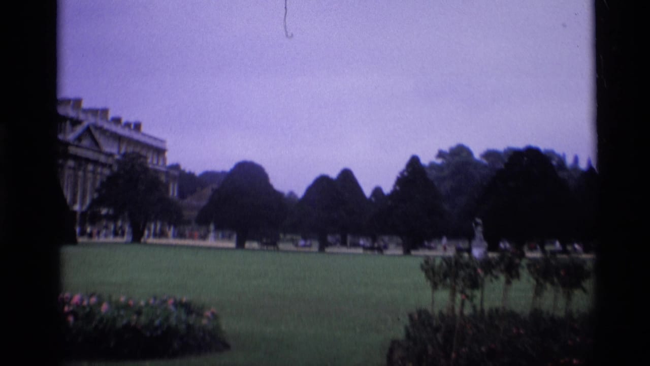 1969 London frame with a sweeping garden lawn, shaped trees, distant visitors, and part of a palace or castle-like building at the left edge.