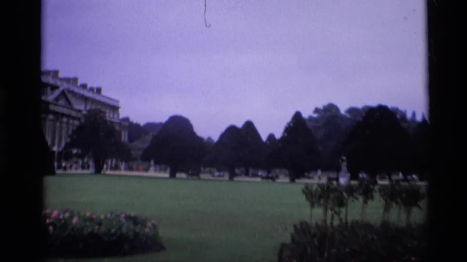 1969 London frame with a sweeping garden lawn, shaped trees, distant visitors, and part of a palace or castle-like building at the left edge.