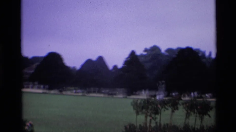 1969 London home-movie frame showing the formal lawn, heavy topiary silhouettes, a distant path, and a small statue or fountain near the trees.
