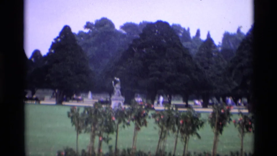 1969 London frame showing a formal garden vista with a white statue, rose-like flowerbeds, broad lawn, mature trees, and distant visitors.