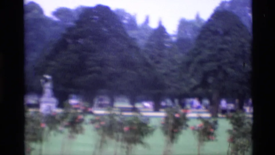 1969 London cemetery grounds filmed in soft 8mm, showing evergreen trees, clipped lawns, flowerbeds, pale edging, and distant stone monuments.