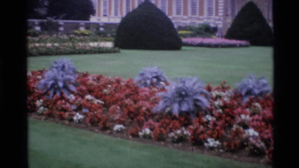 1969 London garden view with red and white flowerbeds, silvery foliage, manicured grass, dark topiary, and a grand building beyond.