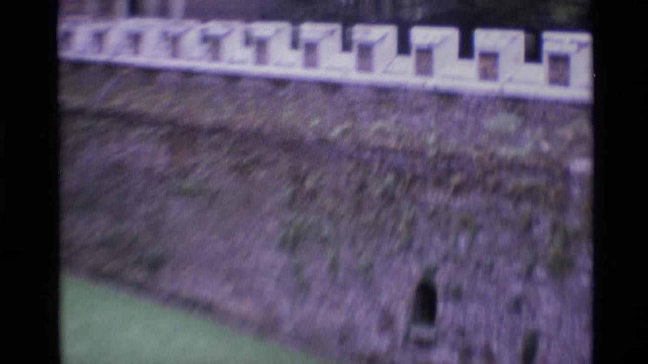 1969 London home-movie frame showing a crenellated stone wall, arched opening, lawn, and soft handheld focus.