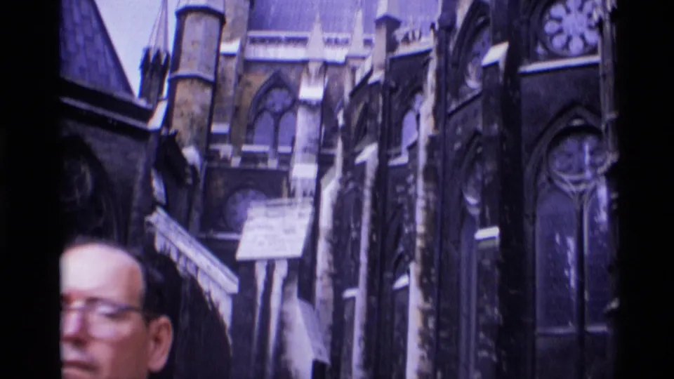 1969 London home-movie frame with a partial adult face crossing the foreground before ornate Gothic cathedral stonework.