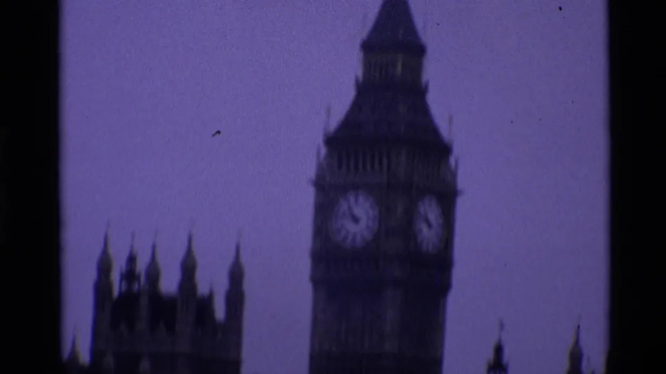 1969 London frame showing Elizabeth Tower and clock faces beside the Palace of Westminster spires in purple-tinted 8mm footage.