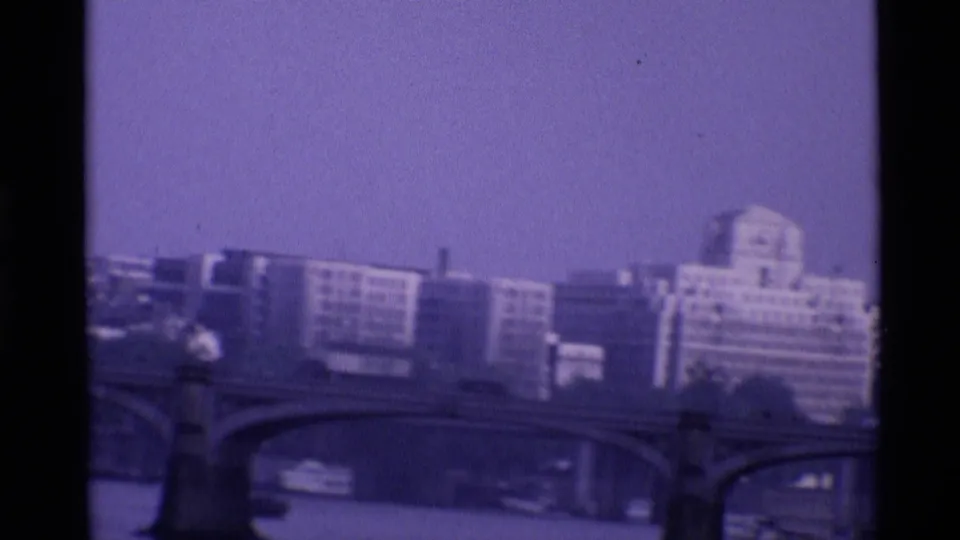 1969 London frame with modern buildings, bridge arches, and river traffic seen through soft purple-tinted 8mm film.