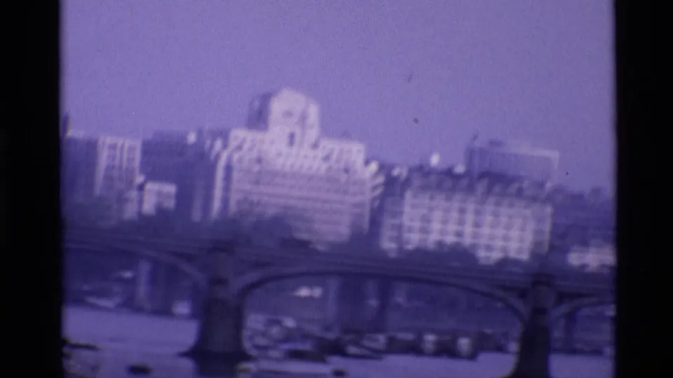 1969 London home-movie frame showing cars on a Thames bridge, boats below, and layered riverside buildings in haze.