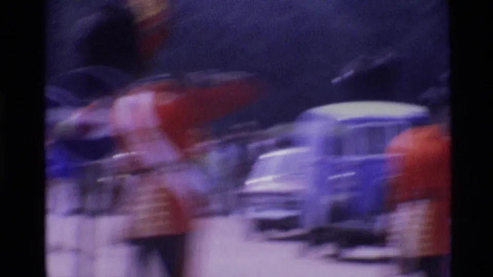 Soft, motion-blurred 1969 London home-movie frame of guards in red uniforms passing a light-colored car on a busy street.