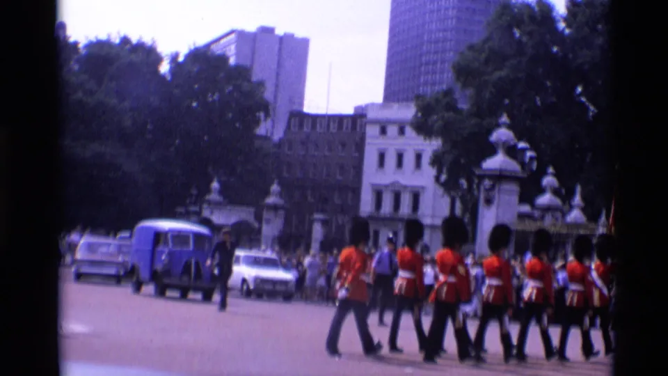 1969 London home-movie frame of guards marching across a broad street with a blue van, cars, onlookers, trees, and buildings behind them.