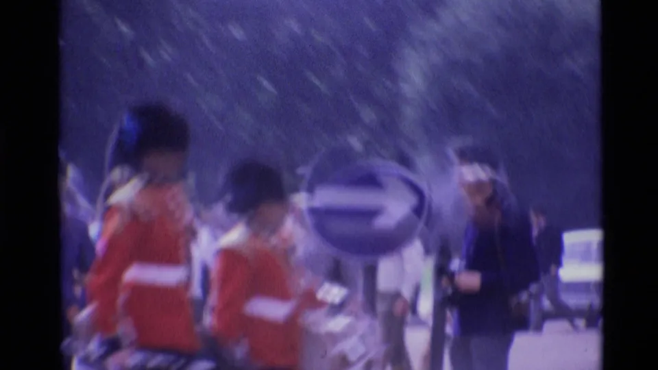 Soft 1969 London street frame with blurred guards in red tunics beside a circular traffic sign and a dark-suited pedestrian.