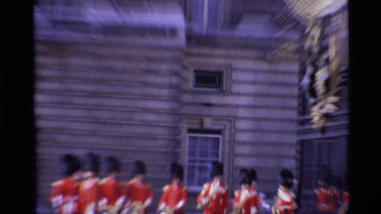 1969 London home-movie frame showing Queen's Guards in red tunics and bearskin hats near a large stone exterior.