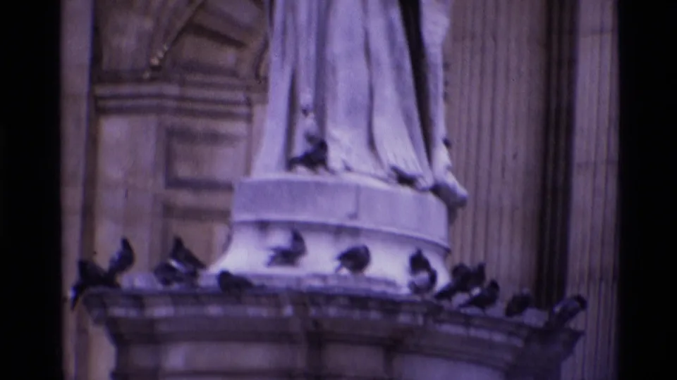 1969 London 8mm frame showing pigeons clustered on a carved stone pedestal beneath a standing statue.