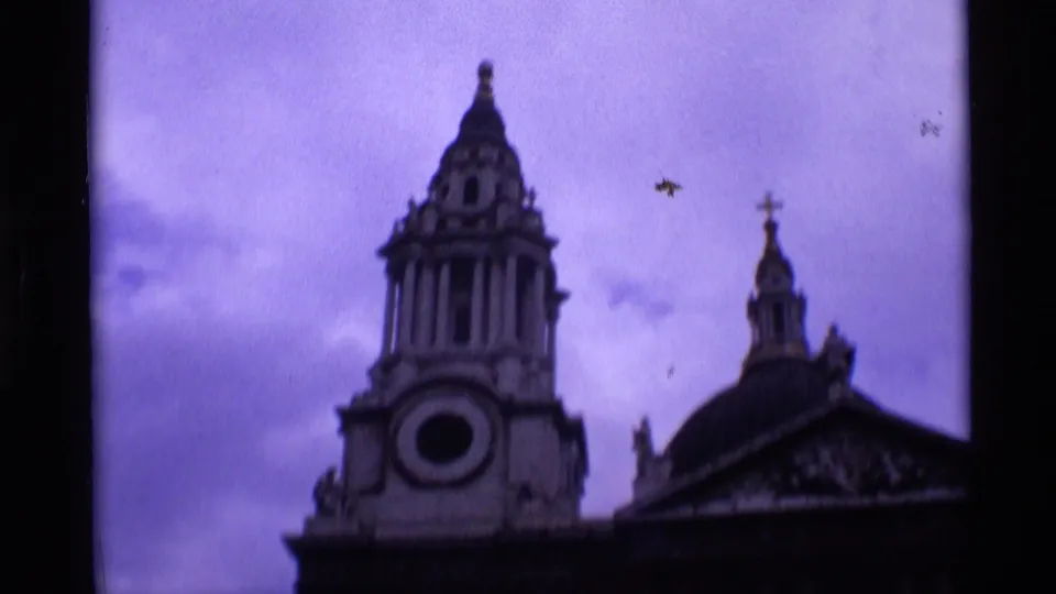 1969 London home-movie frame showing a tower, dome roofline, and cross-topped architectural details against a purple overcast sky.