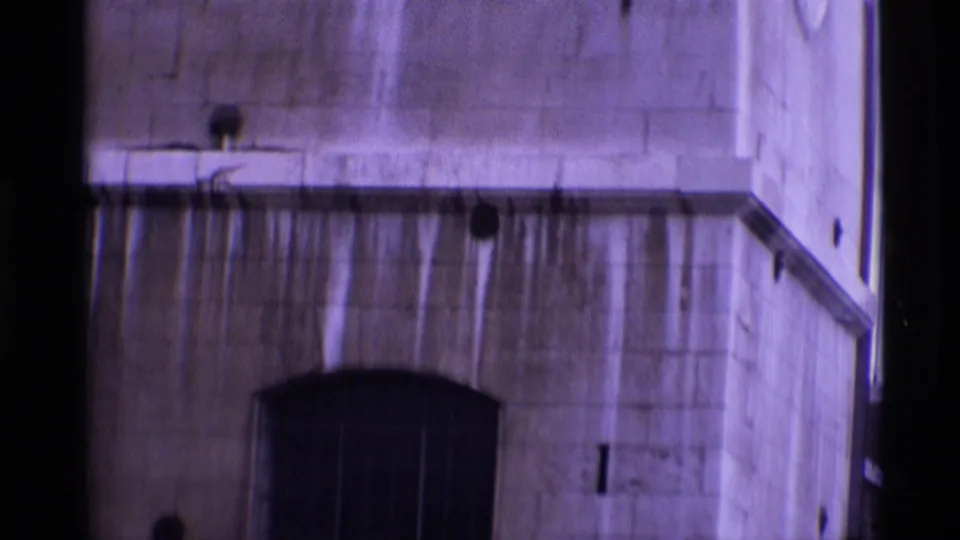 1969 London home-movie frame showing a weathered stone facade with an arched barred doorway and small wall openings.