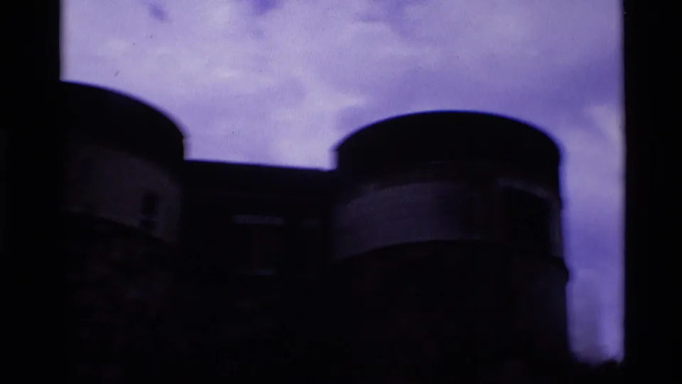 Underexposed 1969 8mm frame showing the Tower of London walls and round towers silhouetted against a purple cloudy sky.