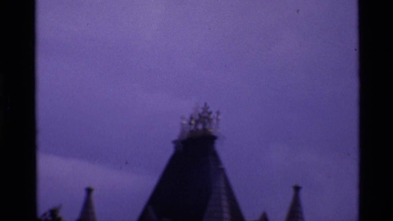 Visitor-shot 8mm view in London of a dark Gothic-style roofline and flanking spires against a purple-shifted sky.