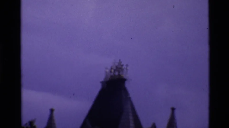 Visitor-shot 8mm view in London of a dark Gothic-style roofline and flanking spires against a purple-shifted sky.